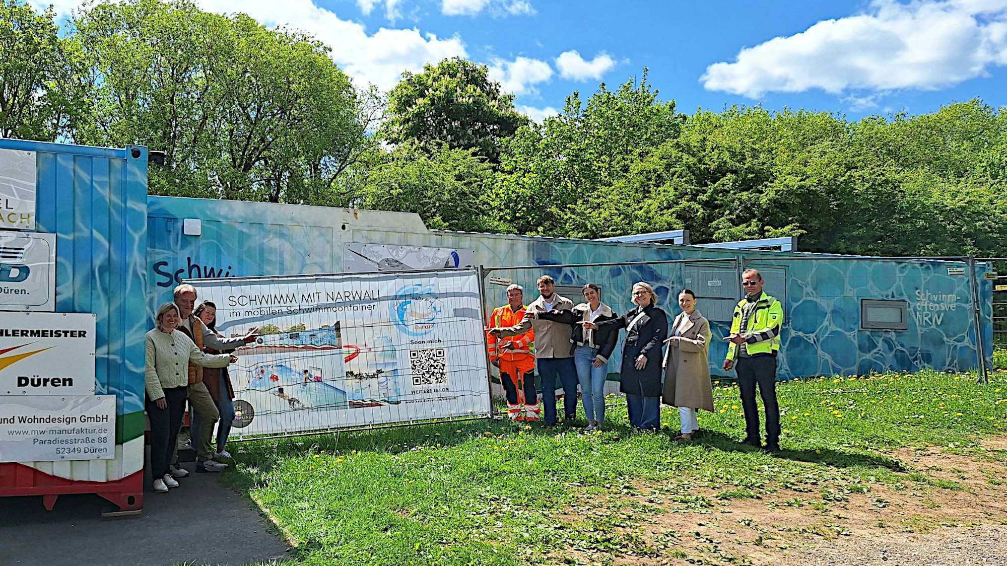 Eine Gruppe Menschen steht auf einer Wiese vor den Containern, in denen sich ein Lehrschwimmbad befindet.