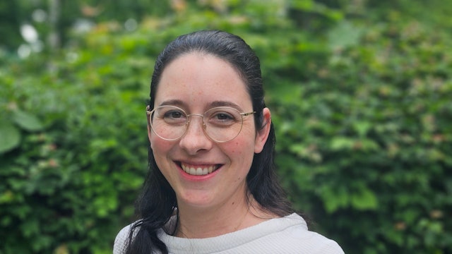 Romina Plonsker hat dunkle Haare und trägt eine runde Brille. Im Hintergrund verschwommenes Grün.