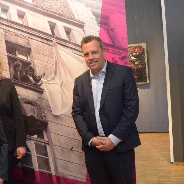 Zwei Frauen und ein Mann stehen vor einem Schwarz-Weiß-Foto. Es zeigt zwei Frauen, die eine weiße Fahne schwenken.
