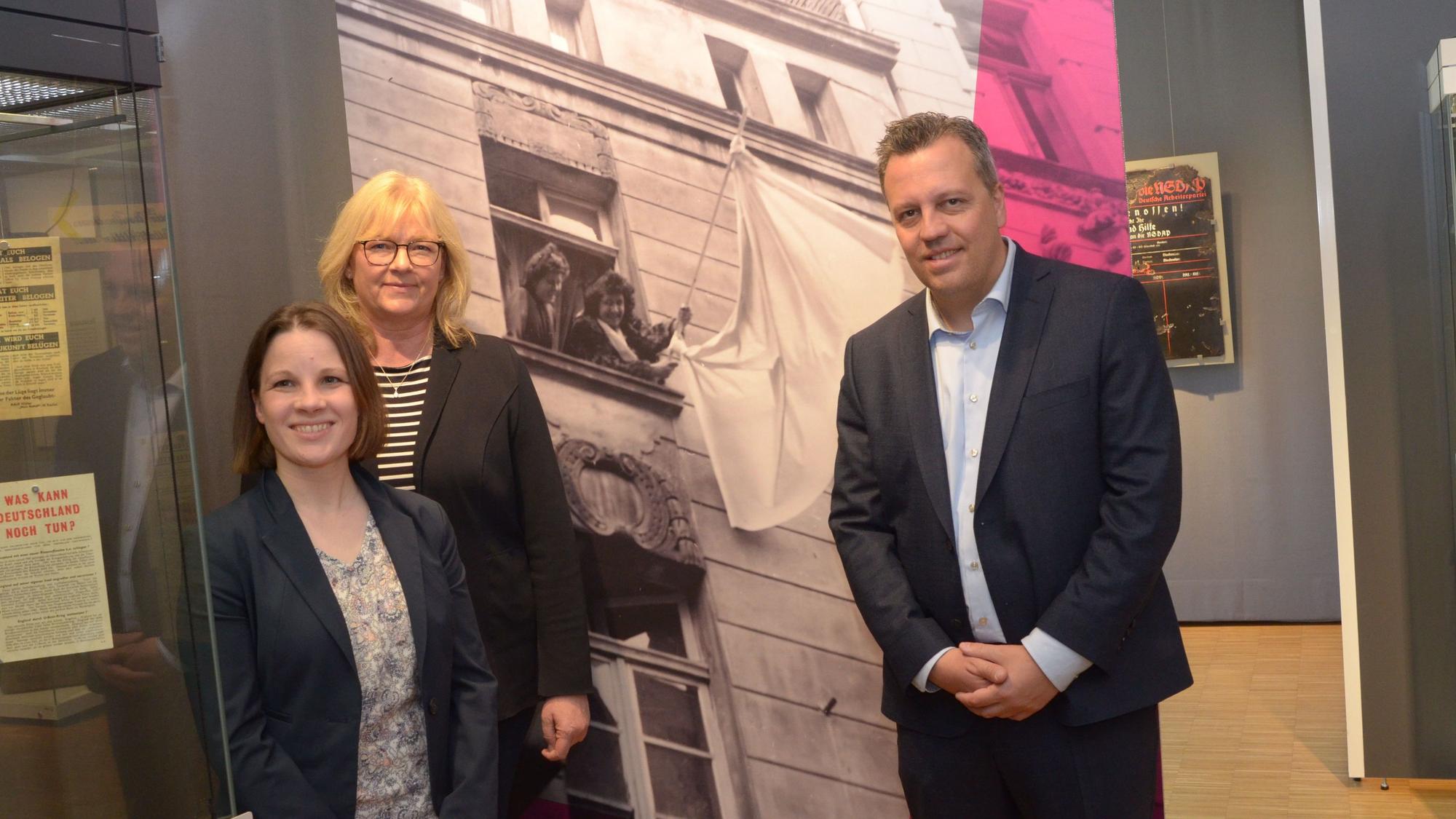 Zwei Frauen und ein Mann stehen vor einem Schwarz-Weiß-Foto. Es zeigt zwei Frauen, die eine weiße Fahne schwenken.