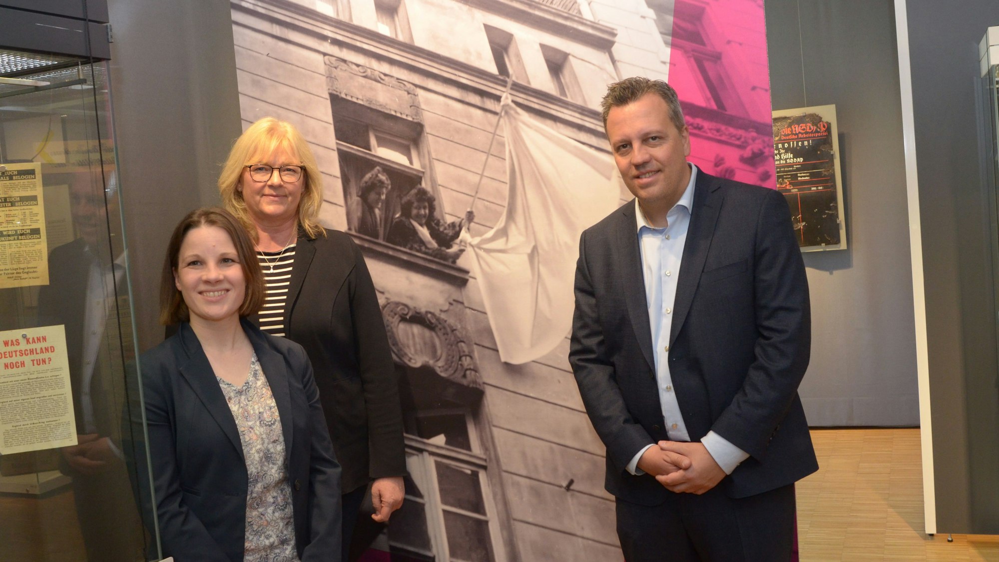 Zwei Frauen und ein Mann stehen vor einem Schwarz-Weiß-Foto. Es zeigt zwei Frauen, die eine weiße Fahne schwenken.