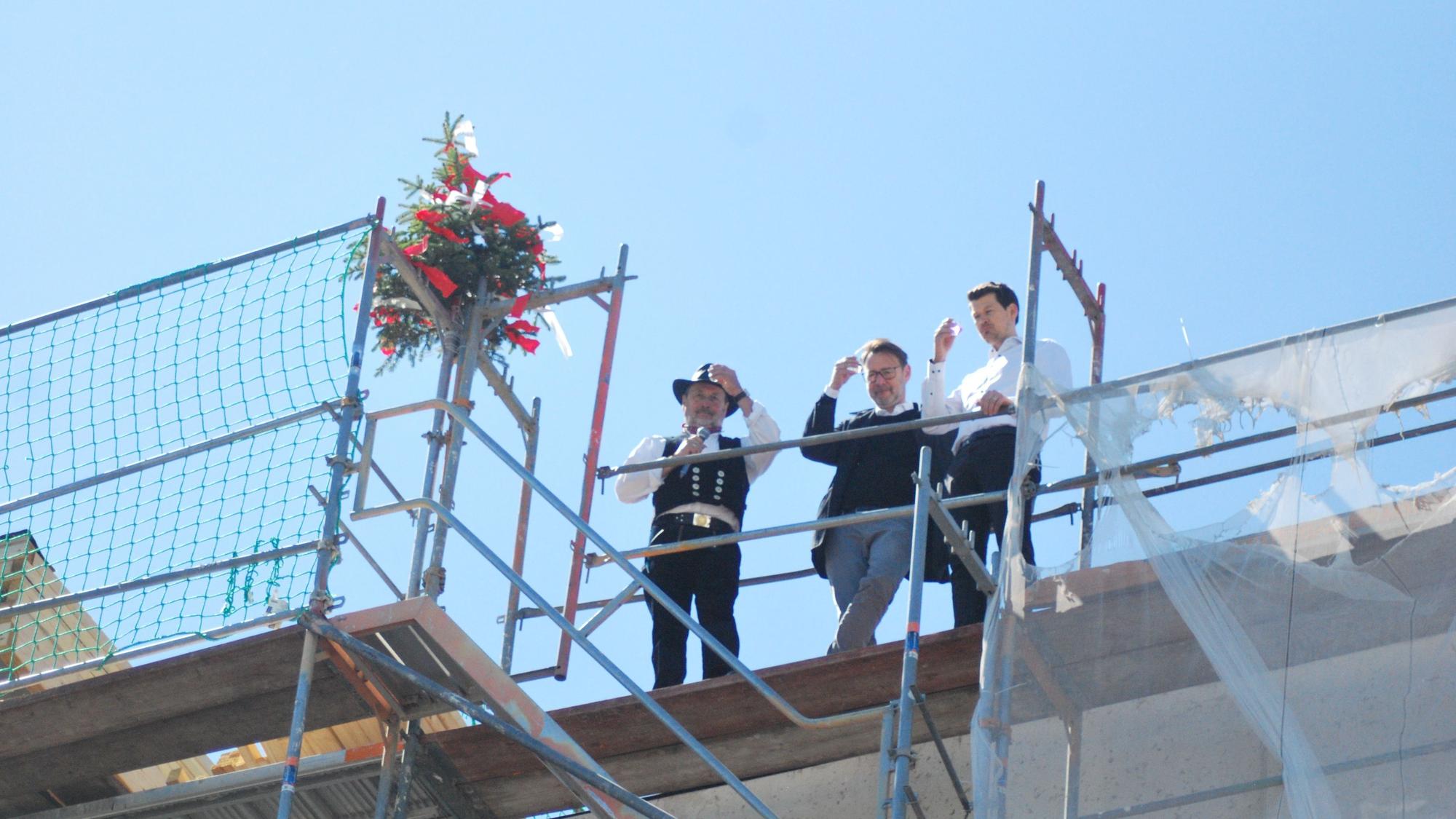 Das Bild zeigt den Zimmermann und zwei weitere Männer auf dem Dach des Neubaus. Sie prosten den Zuschauern zu.