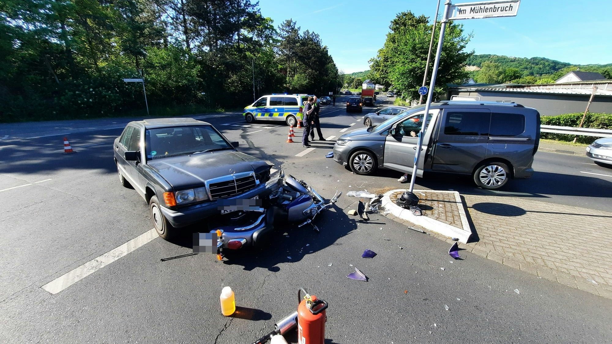 Ein Motorrad, das unter der Frontpartie eines Autos eingeklemmt ist. Im Vordergrund steht ein Feuerlöscher, im Hintergrund stehen Polizisten.