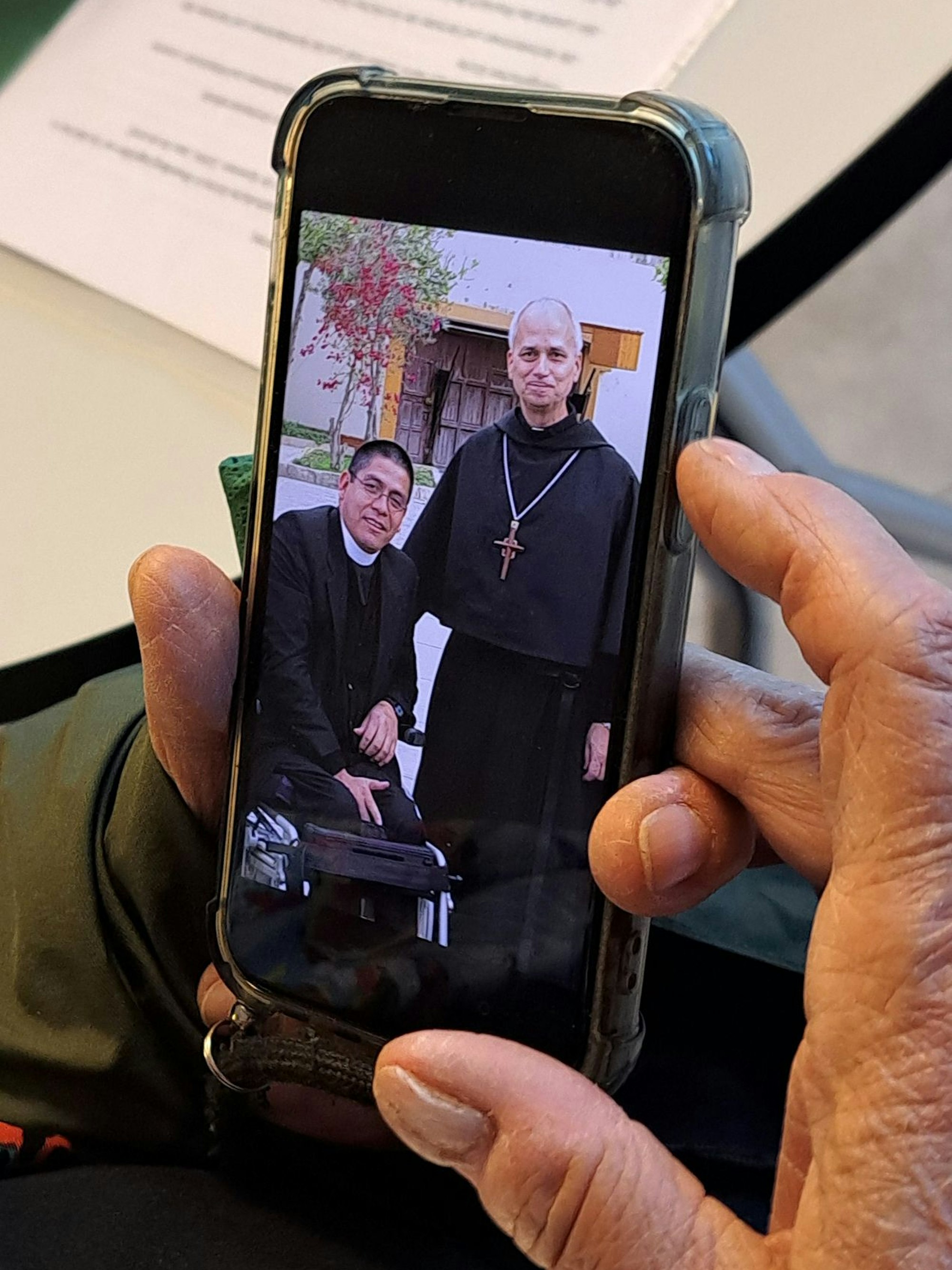 Auf einem Handy in einer Hand sind ein in einem Rollstuhl sitzender Geistlicher und Robert Francis Prevost, der neue Papst Leo XIV., zu sehen.