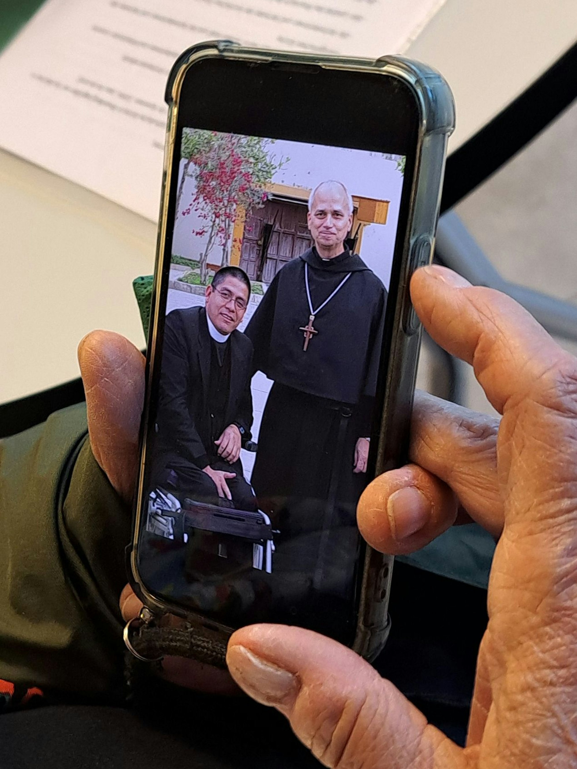 Auf einem Handy in einer Hand sind ein in einem Rollstuhl sitzender Geistlicher und Robert Francis Prevost, der neue Papst Leo XIV., zu sehen.