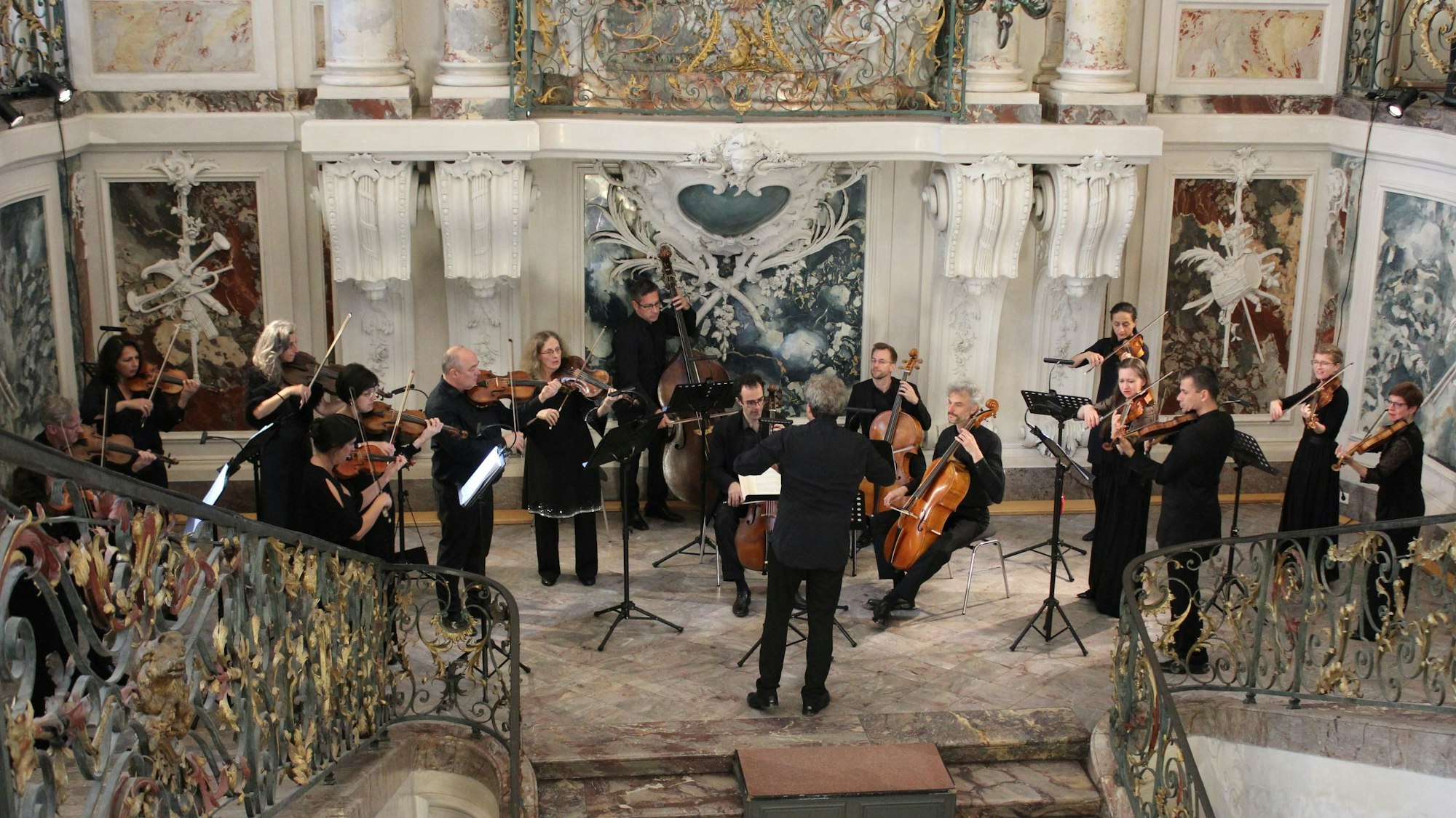 Zu sehen sind Musiker mit unterschiedlichen Instrumenten in einem mit Marmor verkleideten Treppenhaus eines Schlosses.