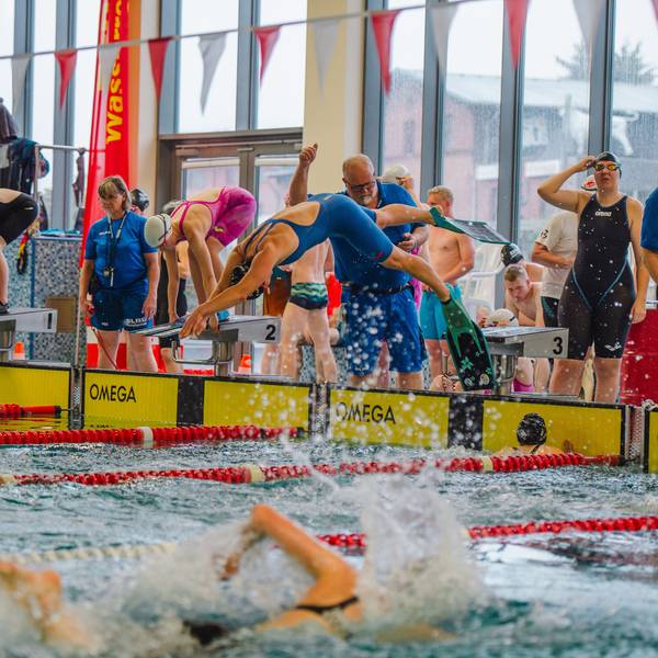 Zu sehen ist ein Schwimmbecken, im Vordergrund ein Schwimmer, im Hintergrund springt einer in das Wasser. Zahlreiche Zuschauer und weitere Schwimmer am Beckenrand sind ebenfalls zu sehen.
