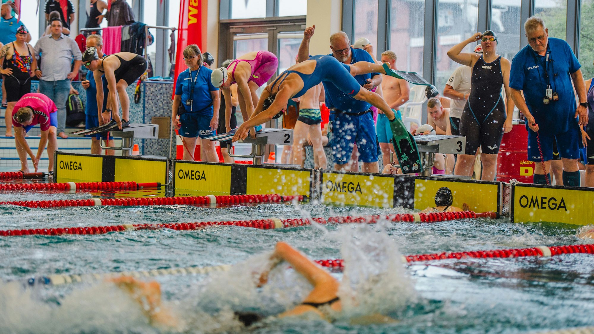 Zu sehen ist ein Schwimmbecken, im Vordergrund ein Schwimmer, im Hintergrund springt einer in das Wasser. Zahlreiche Zuschauer und weitere Schwimmer am Beckenrand sind ebenfalls zu sehen.