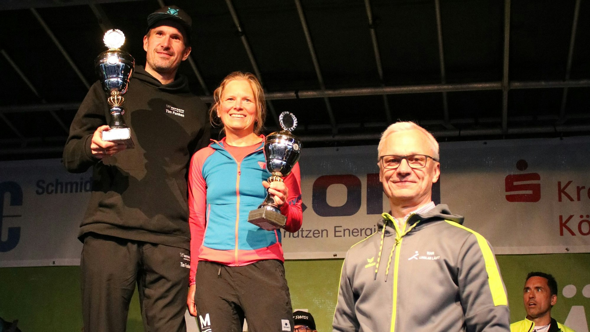 Ein Mann und eine Frau bekommen Pokale von einem zweiten Mann.