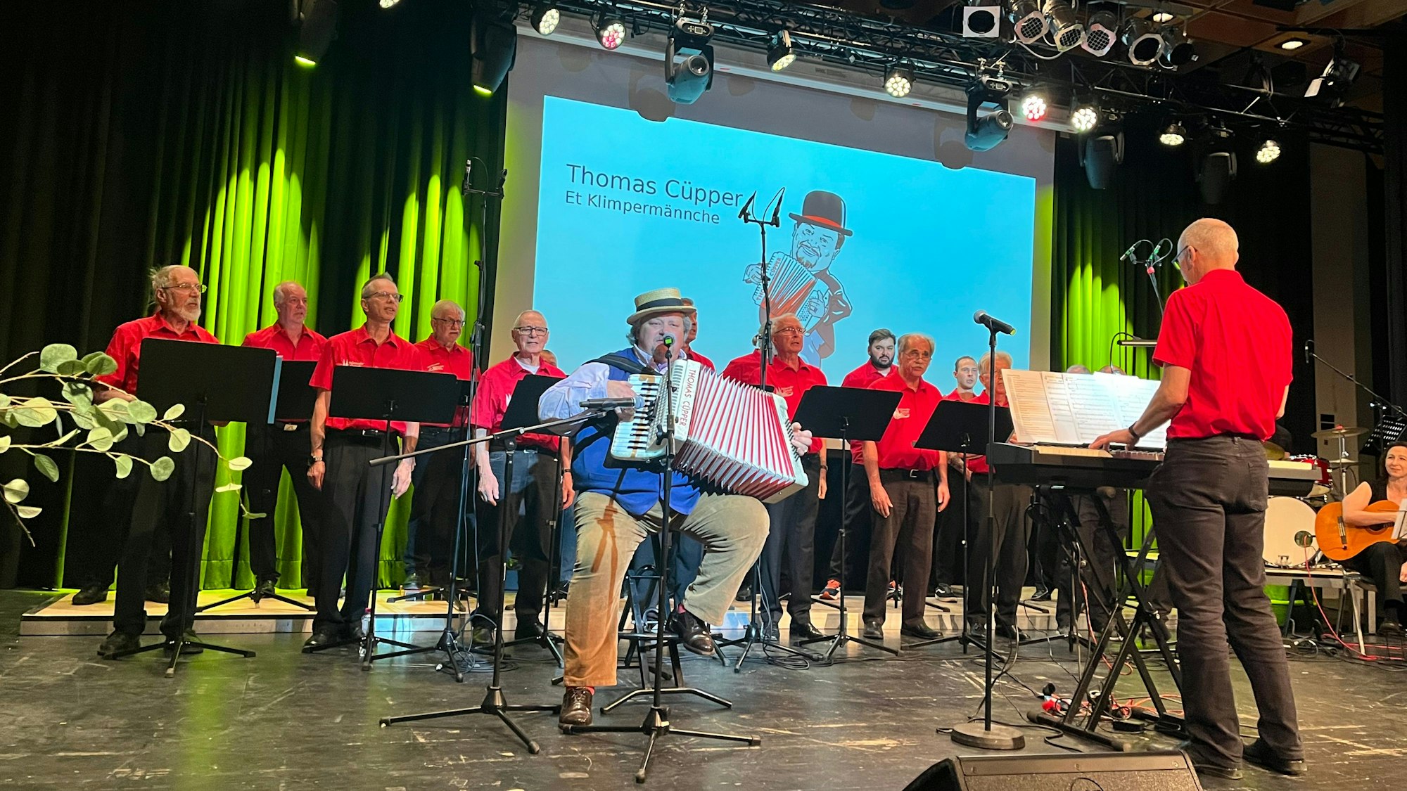 Ältere Herren in schwarzen Hosen und roten Shirts singen auf einer Bühne. Vor ihnen sitzt ein Mann und spielt Akkordeon, ein weiterer Mann spielt Keyboard.