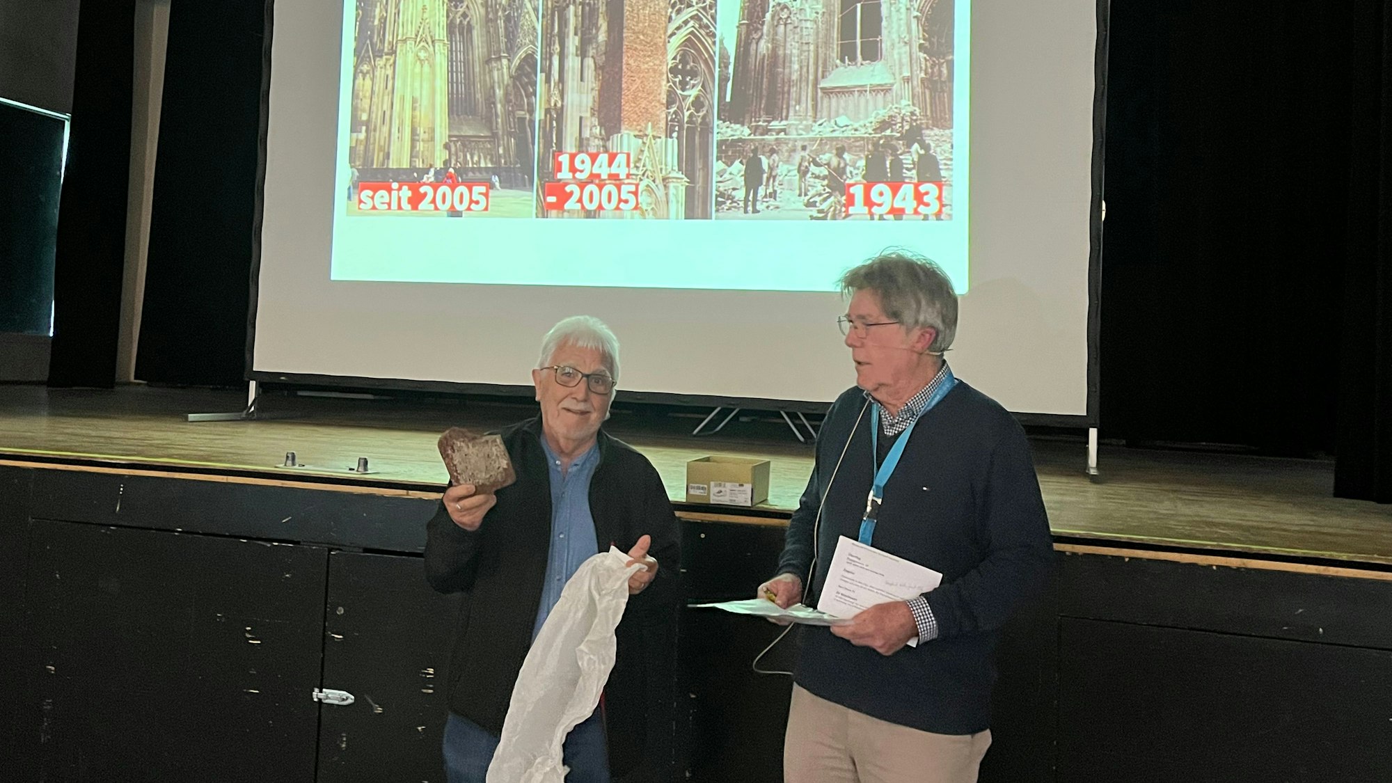 Zwei Männer stehen vor einer Bühne, im Hintergrund ist eine Leinwand mit Bildern vom Dom im Laufe der Zeit zu sehen.