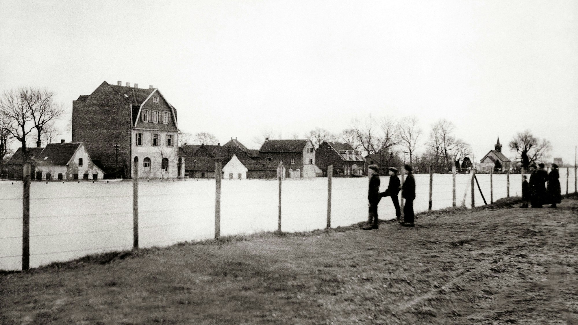 Auf einer Wiese steht eine lange weiße Mauer, dahinter sind Häuser zu sehen.