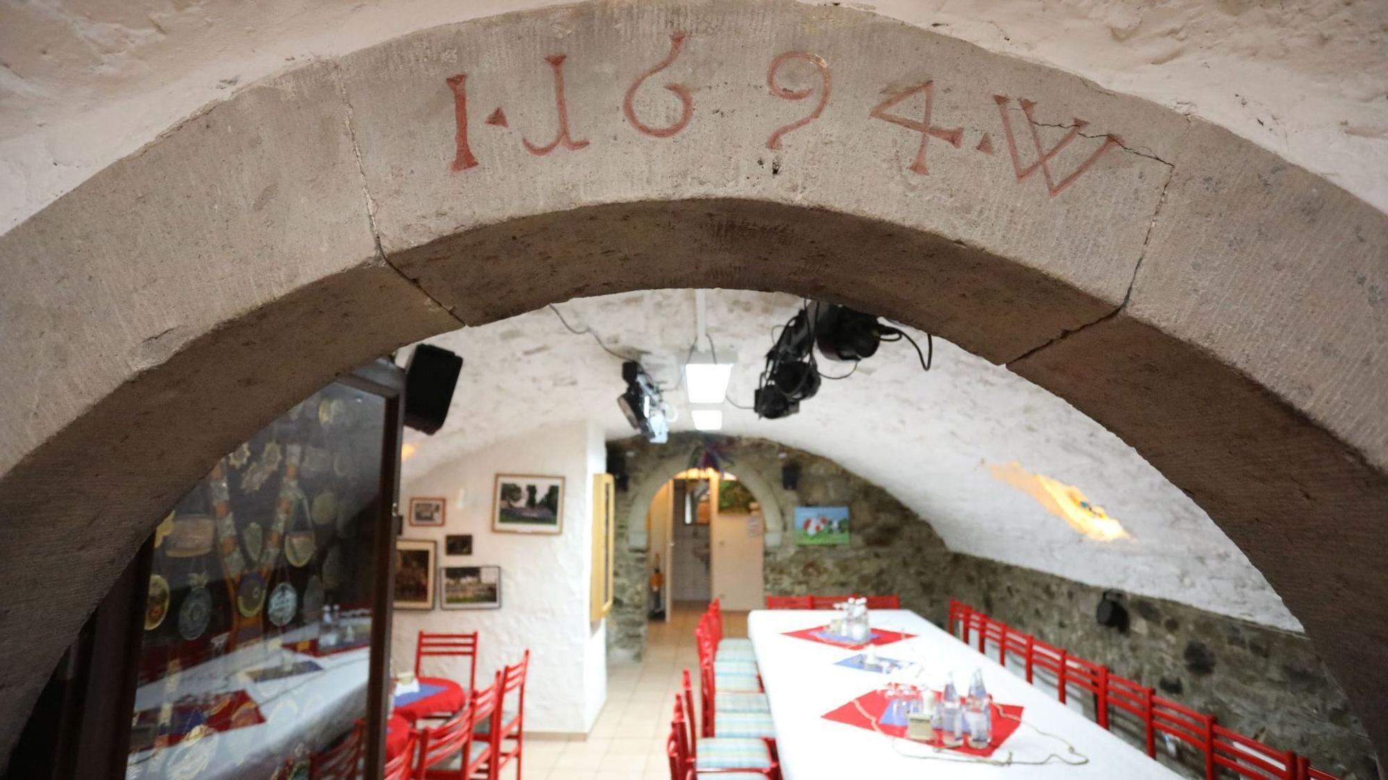 Blick in einen Gewölbekeller, in dem rote Stühle und mit weißen Tischdecken versehene Tische stehen.