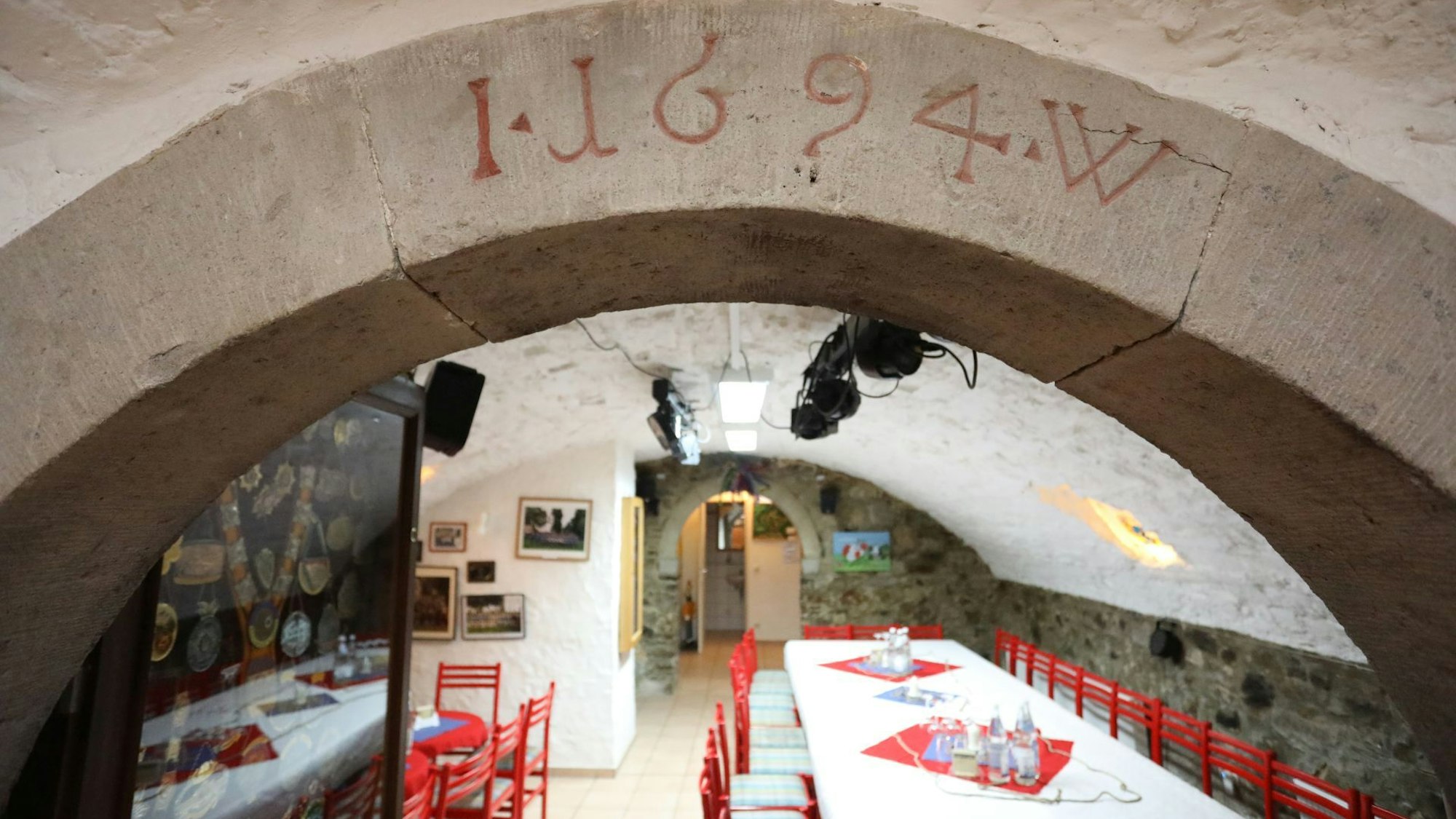 Blick in einen Gewölbekeller, in dem rote Stühle und mit weißen Tischdecken versehene Tische stehen.