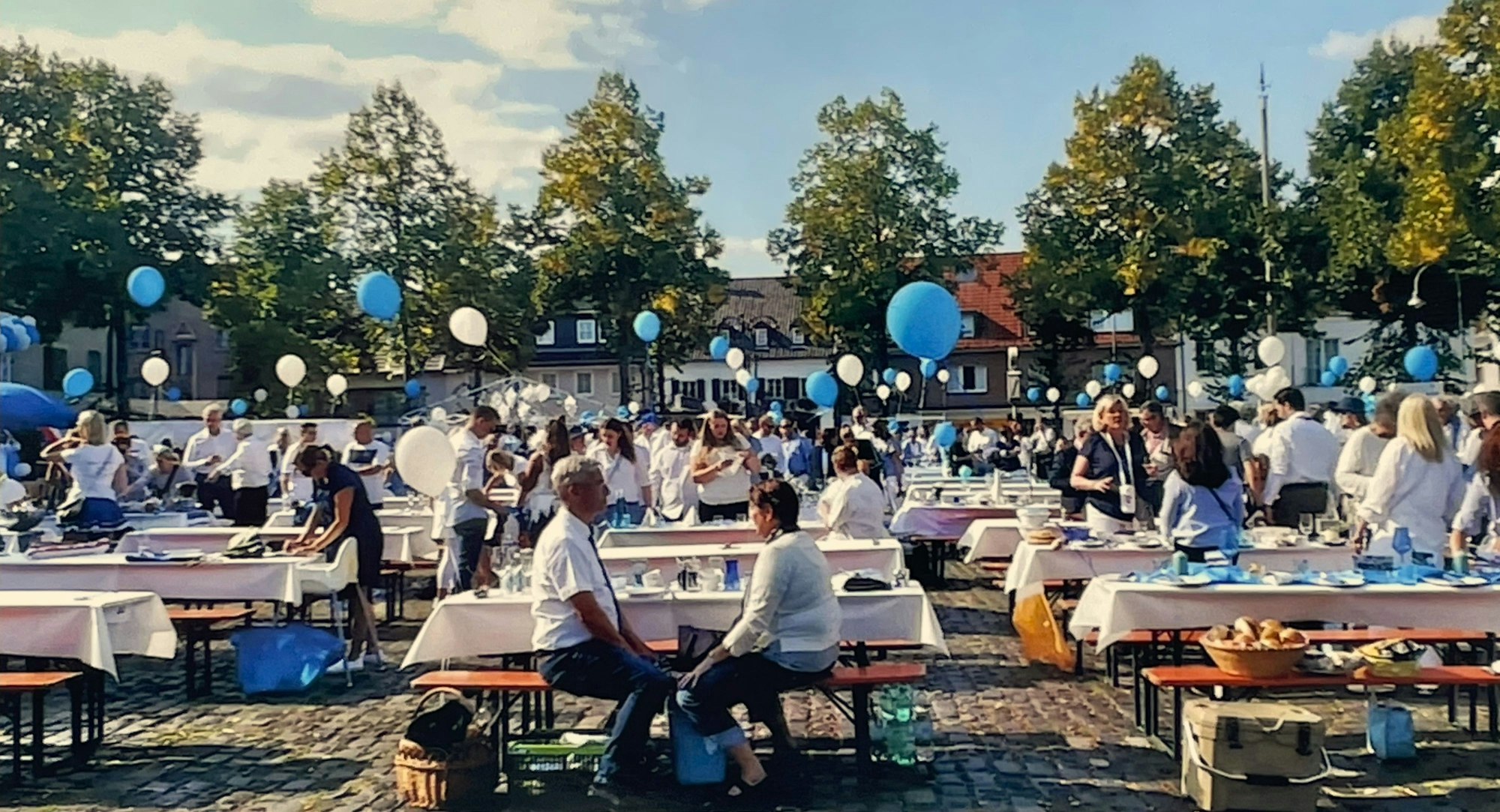 Auf einem Marktplatz stehen zahlreiche Biertische, es gibt Luftbalons in Blau und Weiß, Menschen sitzen an den Tischen.