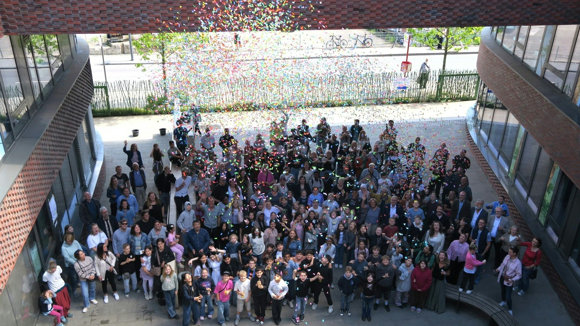 Eine große Menschenmenge steht im Vorhof einer Schule. Auf die Menge regnet es Konfetti.
