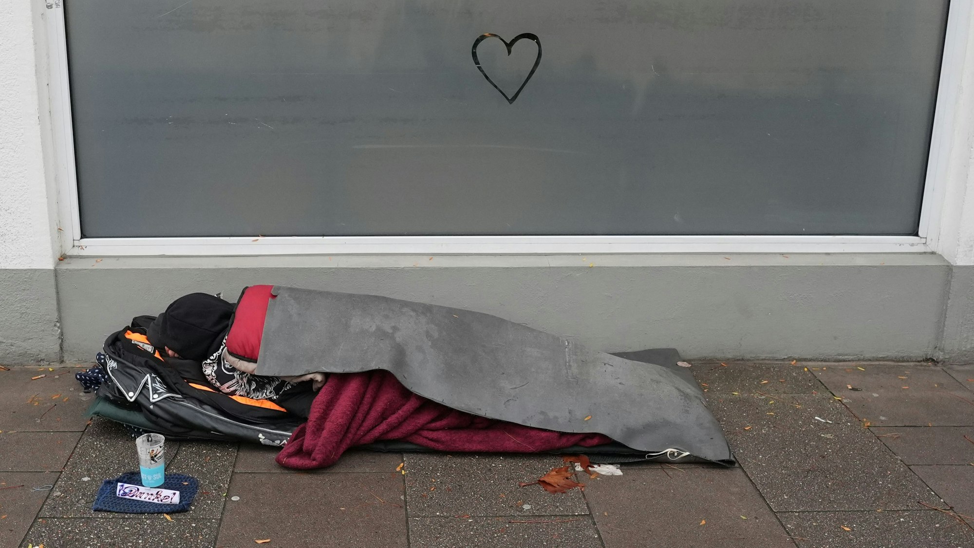 Ein obdachloser Mensch liegt in einem Schlafsack auf dem Gehweg vor einem Supermarkt.