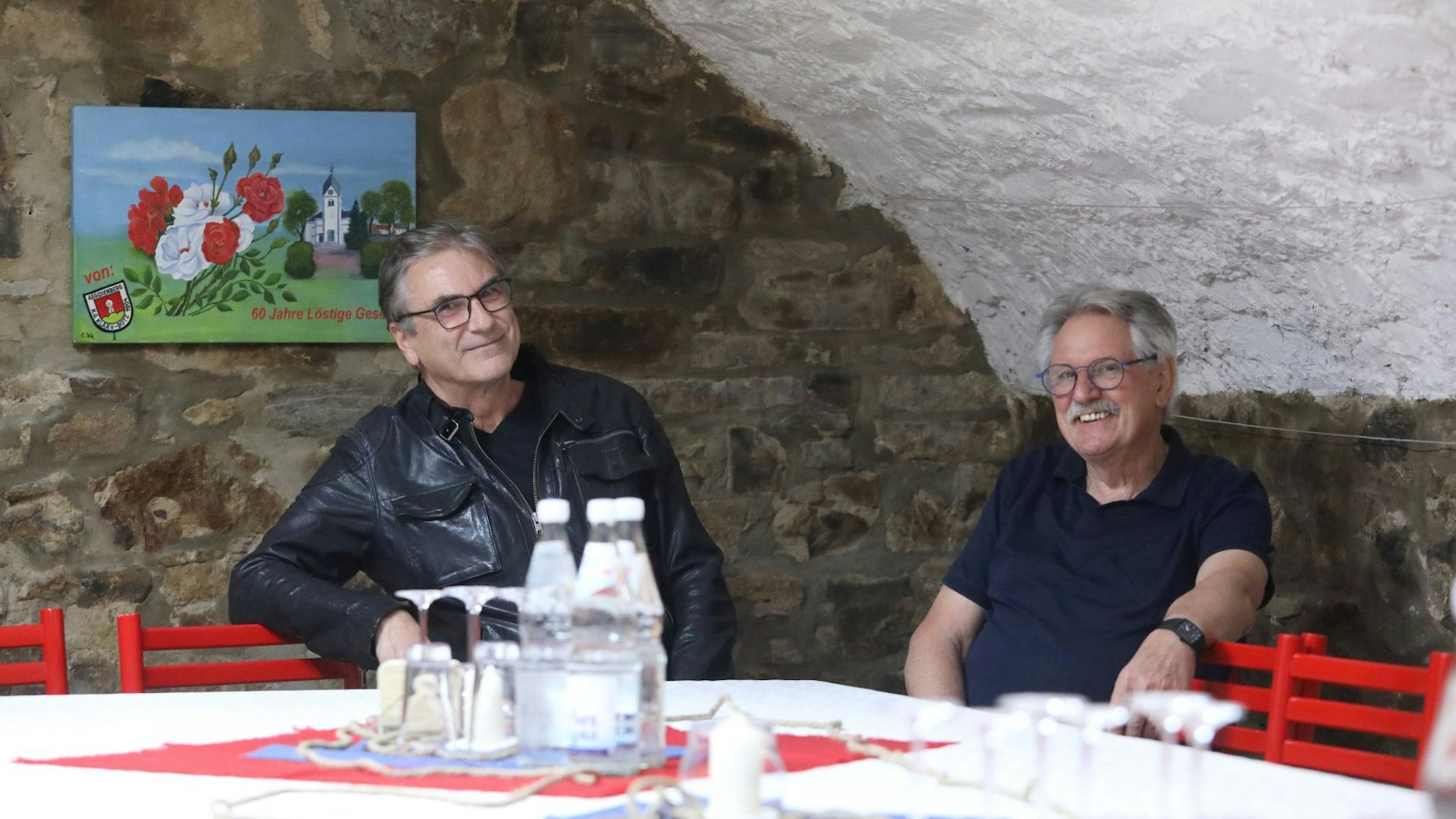Zwei Männer sitzen an einem Tisch, auf dem Gläser und Mineralwasserflaschen stehen.
