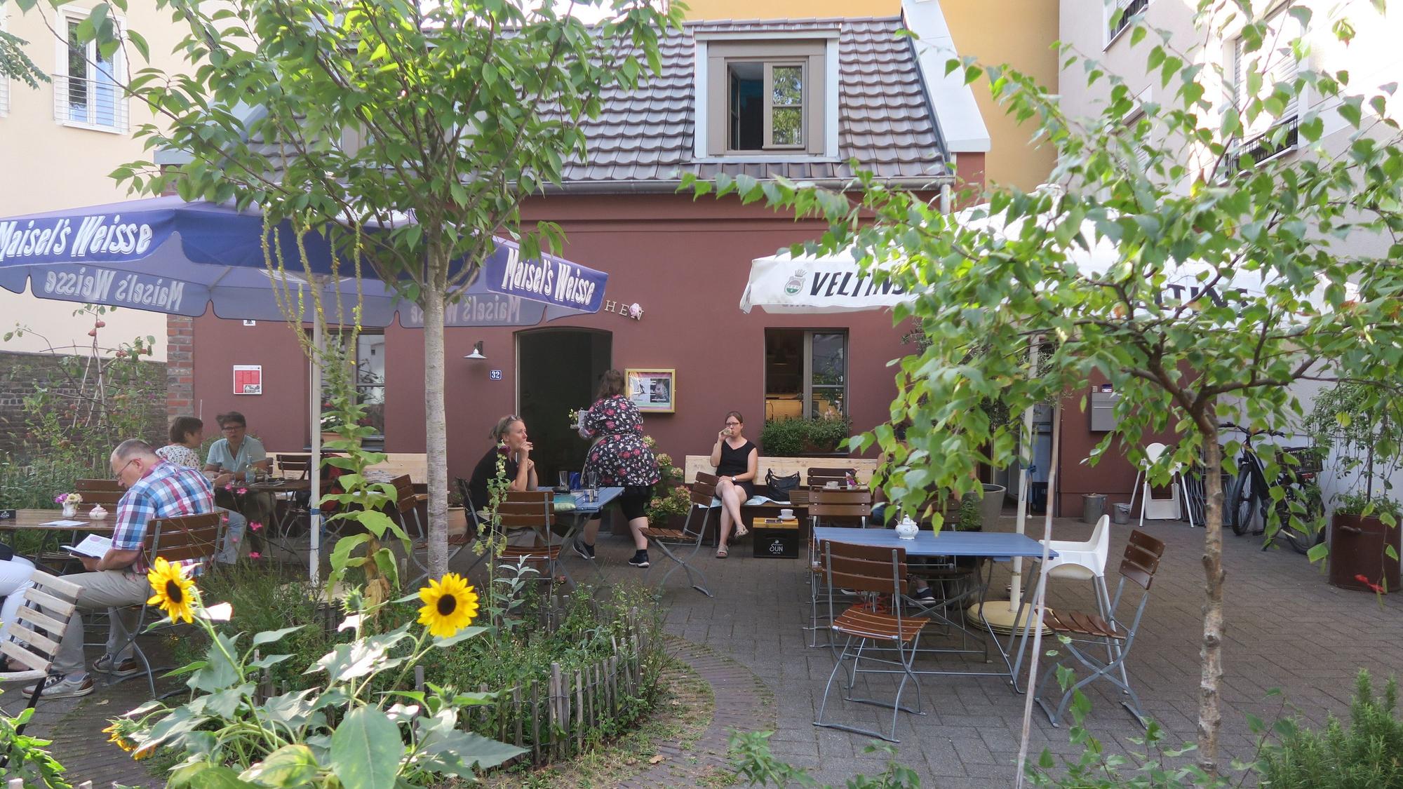 Café-Tische stehen in einem von Pflanzen und einem Holzzaun umrahmten Platz vor einem roten Häuschen.