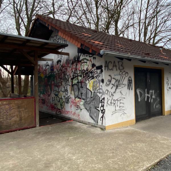 Man sieht ein kleines Haus, das mit vielen Schmierereien überzogen ist und an der linken Seite einen hölzernen Anbau hat.