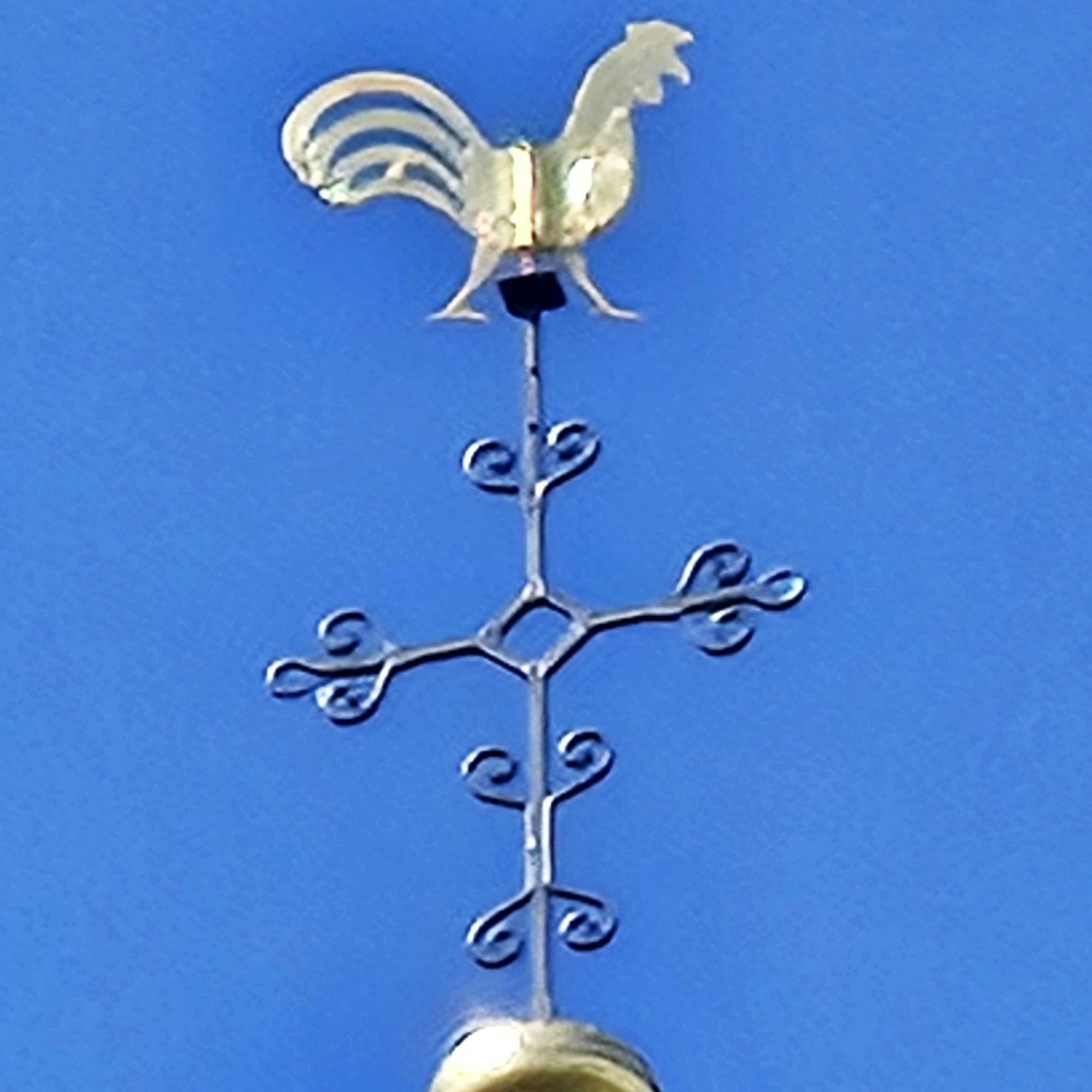 Ein goldener Hahn thront über einem stilisierten Kreuz.