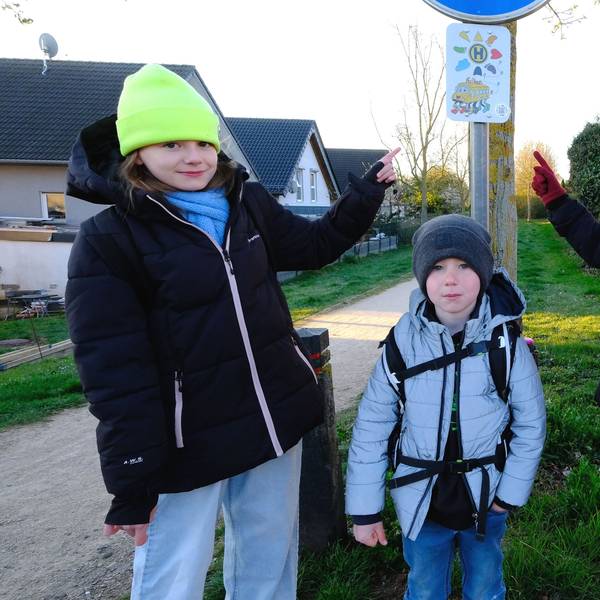 Vier Grundschulkinder, drei Mädchen in schwwarzen Jacken und ein kleinerer Junge, an der Fußbus-Haltestelle in Mechernich-Nord. Zwei Mädchen deuten auf das Haltestellen-Schild.