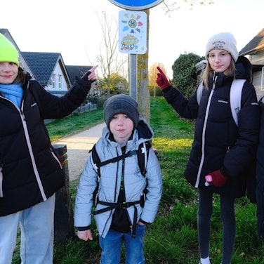 Vier Grundschulkinder, drei Mädchen in schwwarzen Jacken und ein kleinerer Junge, an der Fußbus-Haltestelle in Mechernich-Nord. Zwei Mädchen deuten auf das Haltestellen-Schild.