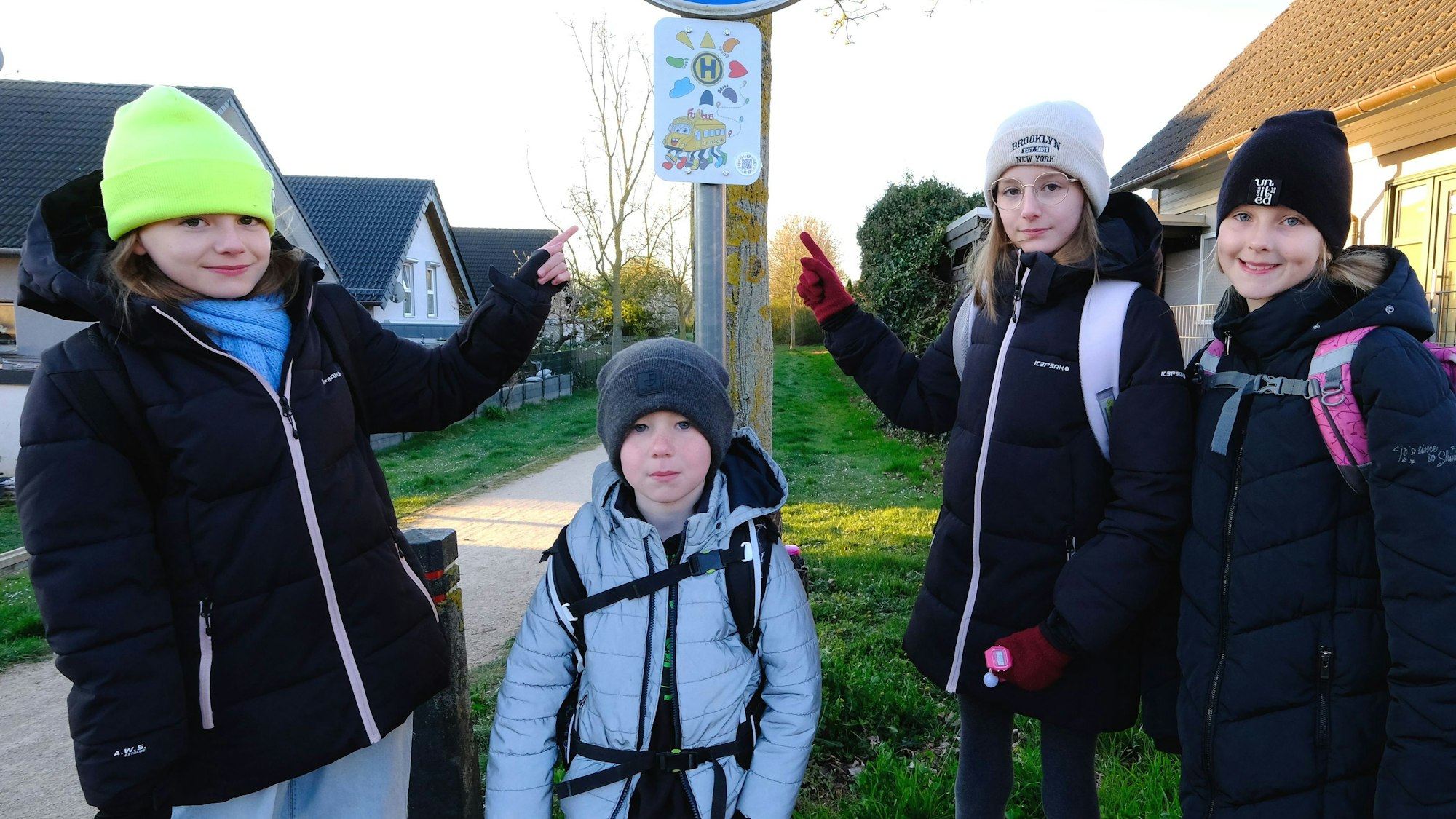 Vier Grundschulkinder, drei Mädchen in schwwarzen Jacken und ein kleinerer Junge, an der Fußbus-Haltestelle in Mechernich-Nord. Zwei Mädchen deuten auf das Haltestellen-Schild.