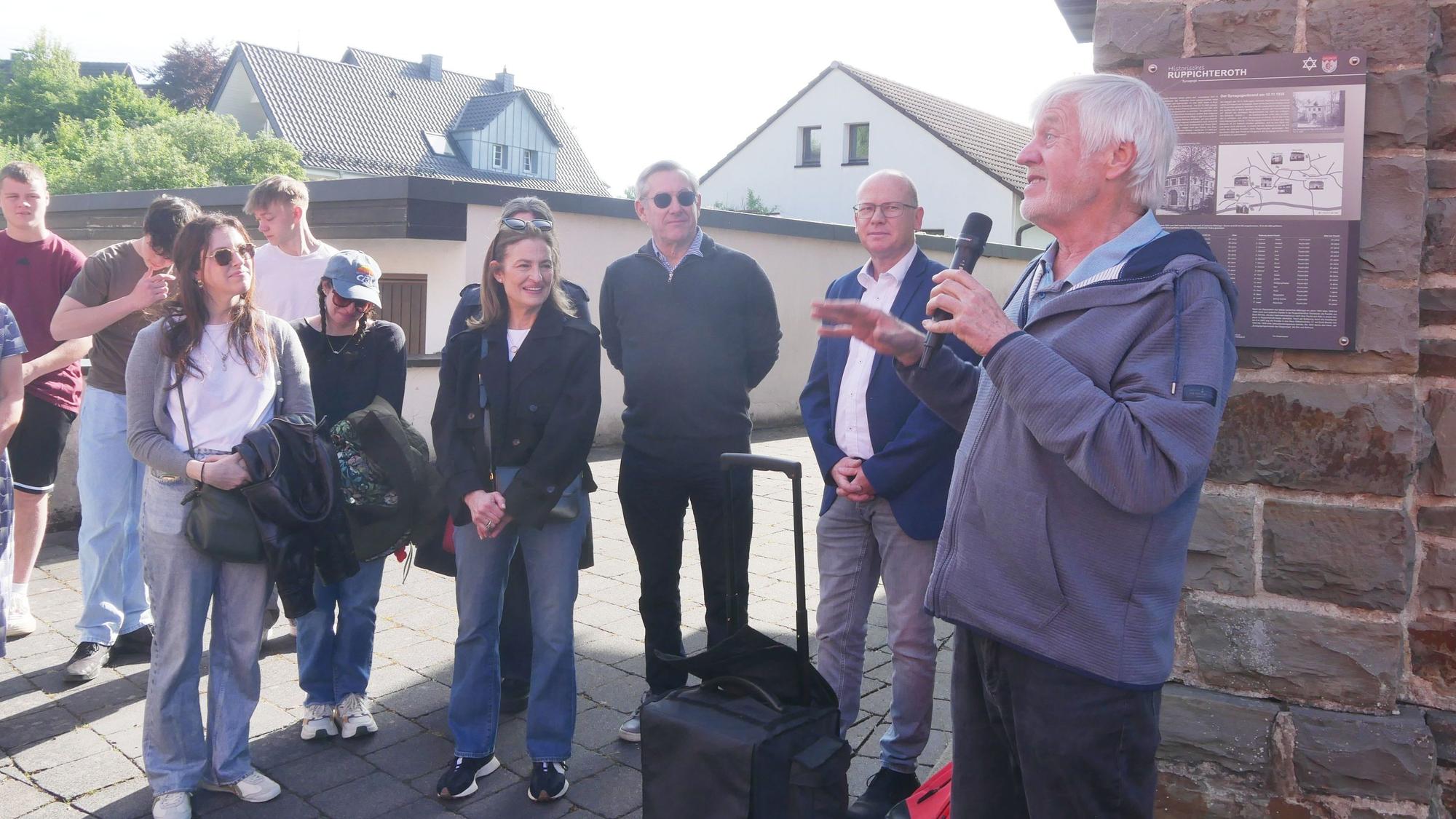 Ein Mann spricht im Freien in ein Mikrofon; einige Personen hören ihm zu.