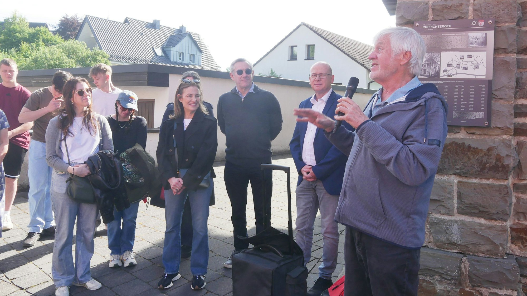 Ein Mann spricht im Freien in ein Mikrofon; einige Personen hören ihm zu.