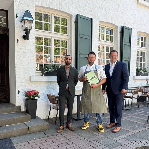 Drei Männer stehen vor einem Restaurant, der in der Mitte hält eine Speisekarte mit dem Schriftzug „Leon“ in den Händen.