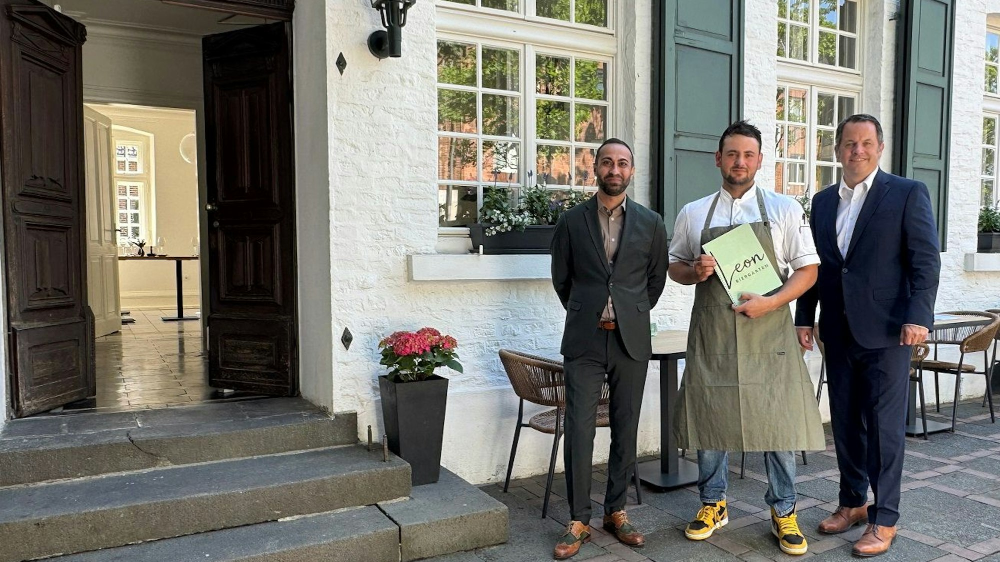 Drei Männer stehen vor einem Restaurant, der in der Mitte hält eine Speisekarte mit dem Schriftzug „Leon“ in den Händen.