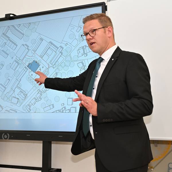 Rolko steht vor einer Leinwand, auf der der Lageplan für den Neubau abgebildet ist.