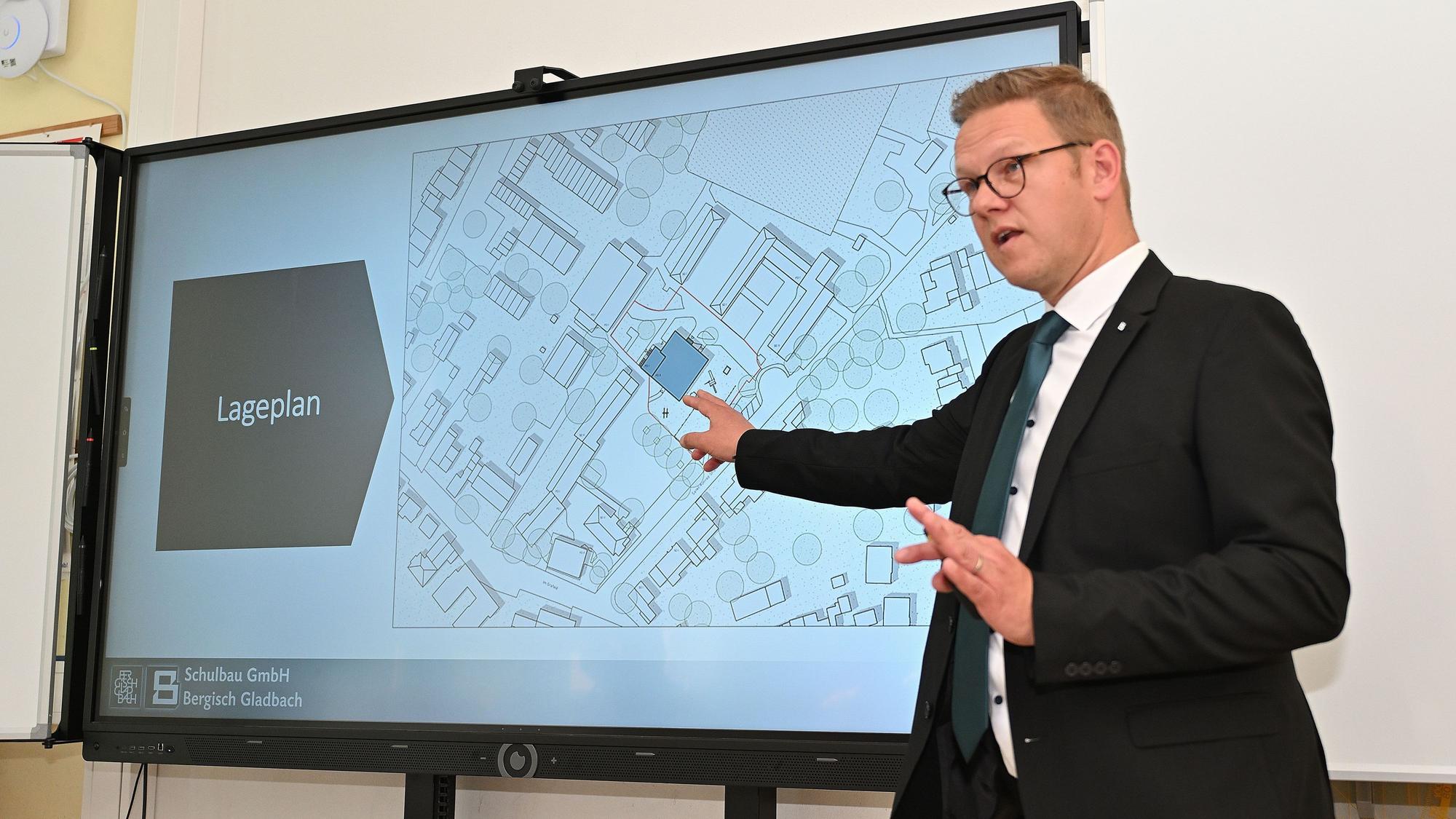 Rolko steht vor einer Leinwand, auf der der Lageplan für den Neubau abgebildet ist.
