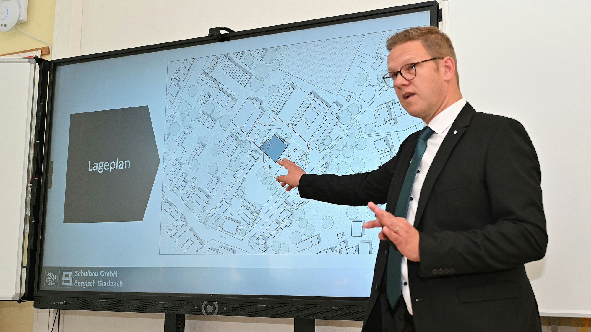 Rolko steht vor einer Leinwand, auf der der Lageplan für den Neubau abgebildet ist.