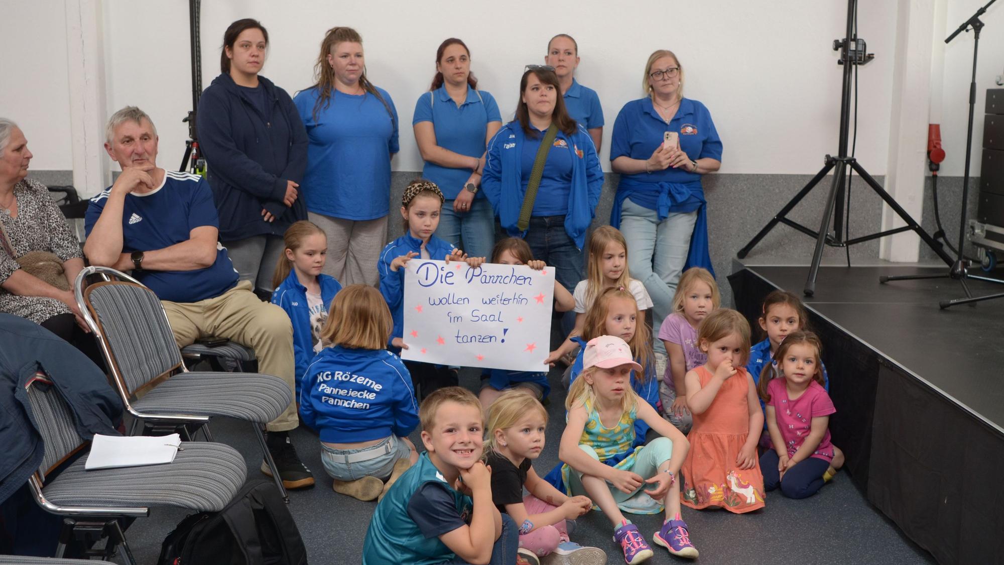 Das Bild zeigt Kinder und ihre Betreuerinnen, einige davon in blauen Jacken und T-Shirts. Drei Mädchen halten ein Plakat in die Höhe mit der Aufschrift „Die Pännchen wollen weiterhin im Saal tanzen“.