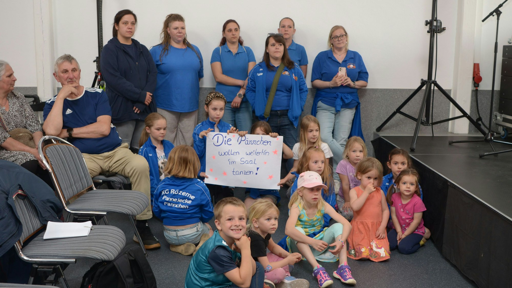 Das Bild zeigt Kinder und ihre Betreuerinnen, einige davon in blauen Jacken und T-Shirts. Drei Mädchen halten ein Plakat in die Höhe mit der Aufschrift „Die Pännchen wollen weiterhin im Saal tanzen“.