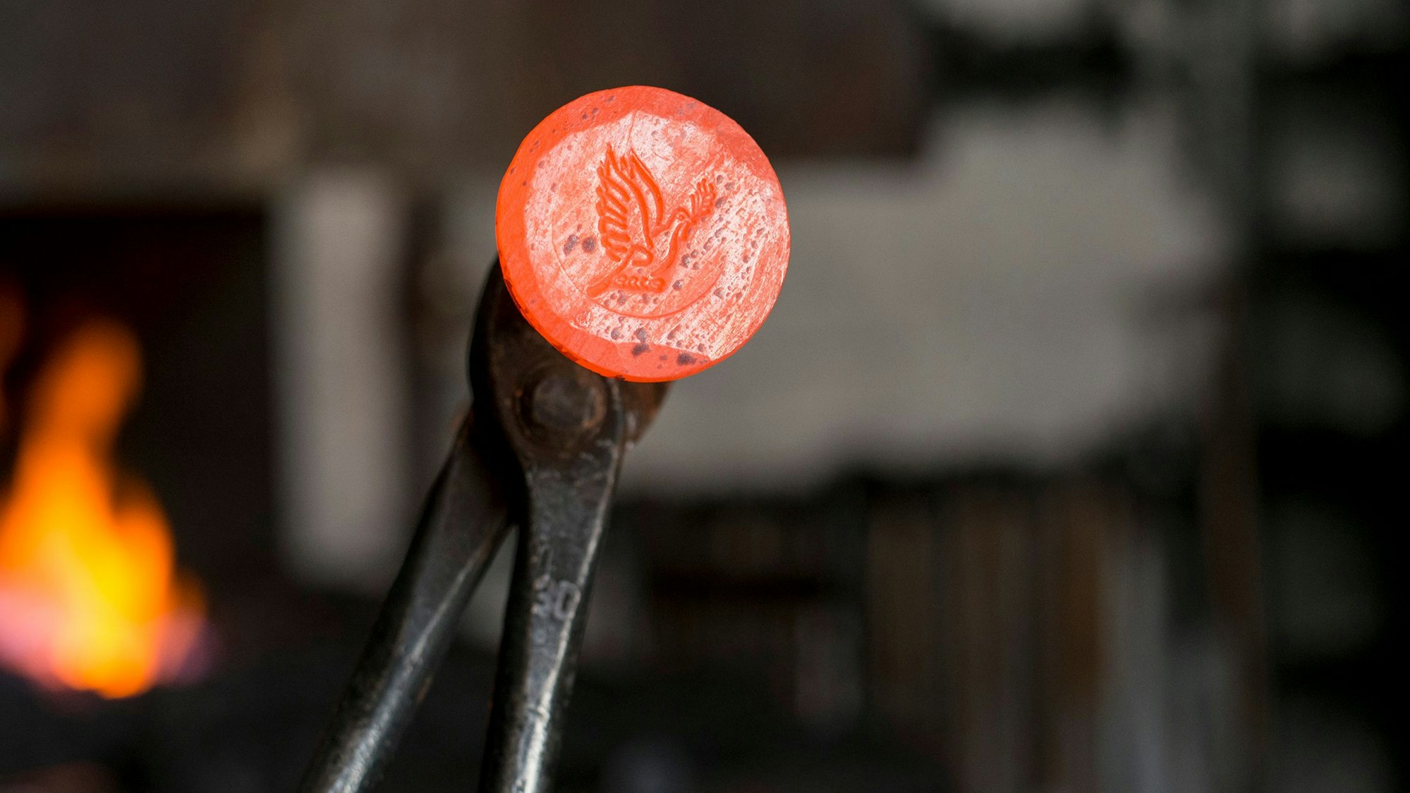 Ein glühender Friedensnagel wird mit einer Zange in die Höhe gehalten, im Hintergrund ist eine Schmiedeesse zu sehen.