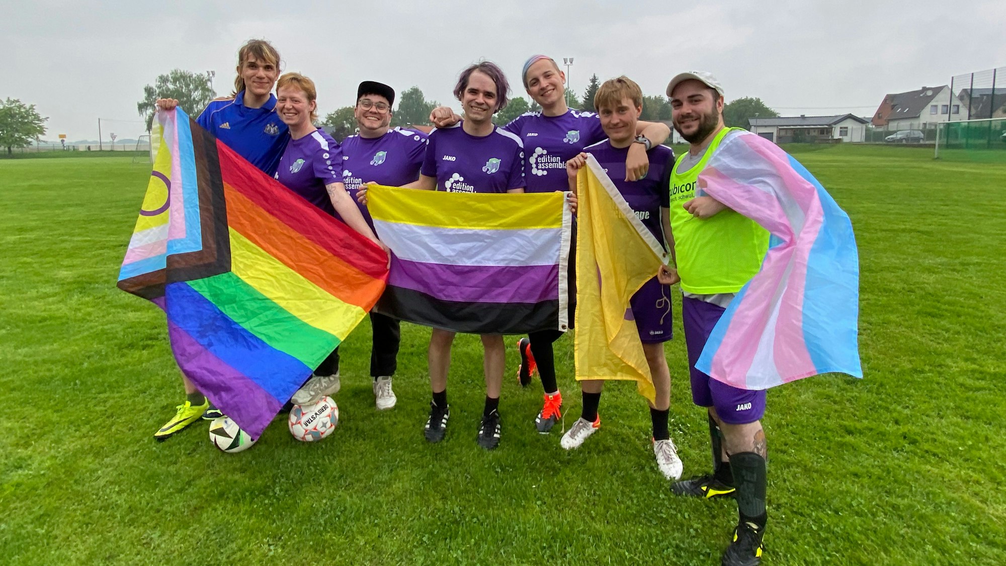 Sieben Personen der queeren Fußball-Truppe stehen Arm in Arm zusammen und halten verschiedene LGBTQIA-Fahnen fest.
