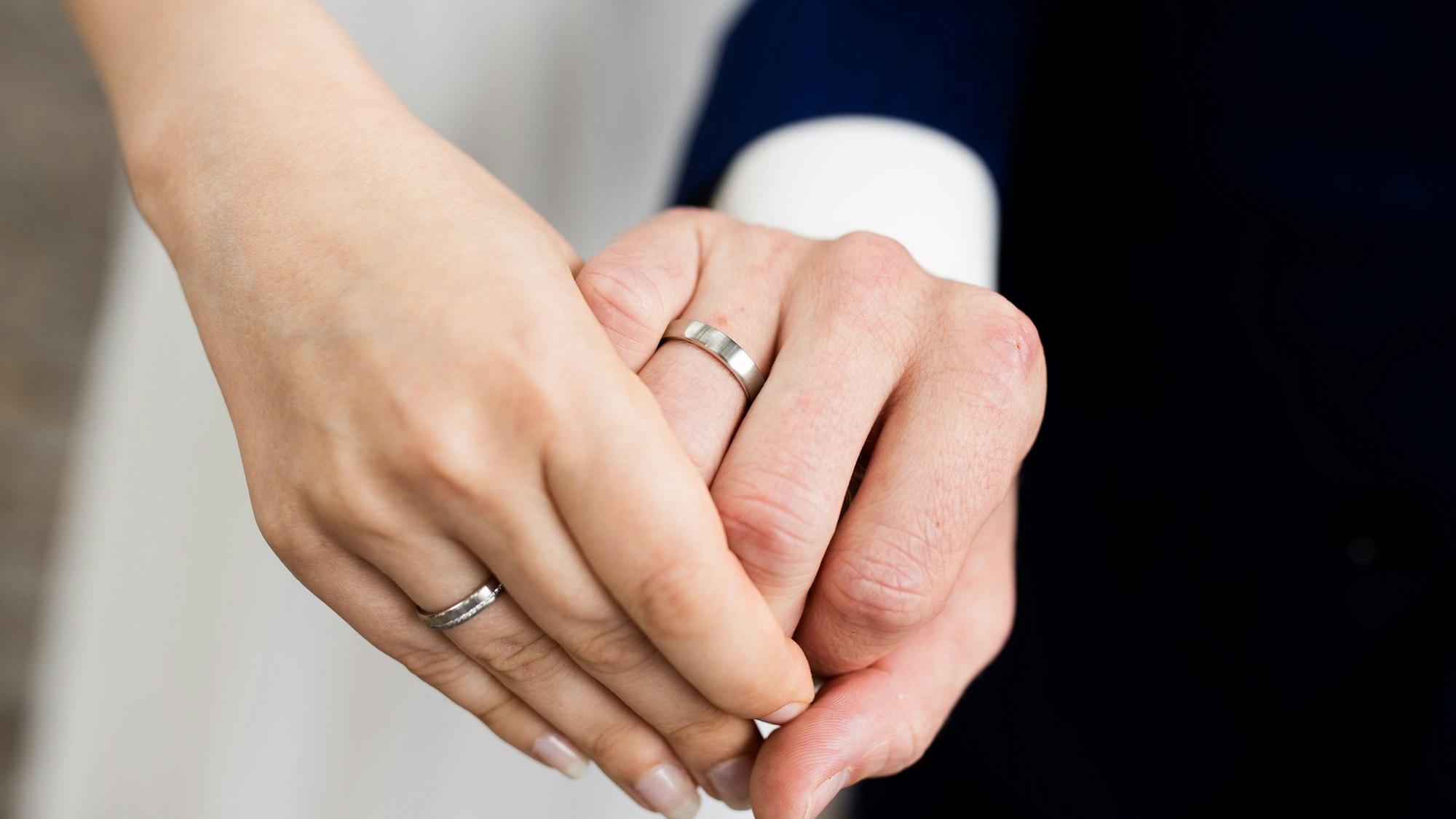 Auf dem Bild sieht man zwei Hände. die sich halten und beide einen Eheringen tragen. Im Hintergrund erkennt man ein Hochzeitskleid und einen schwarzen Anzug.