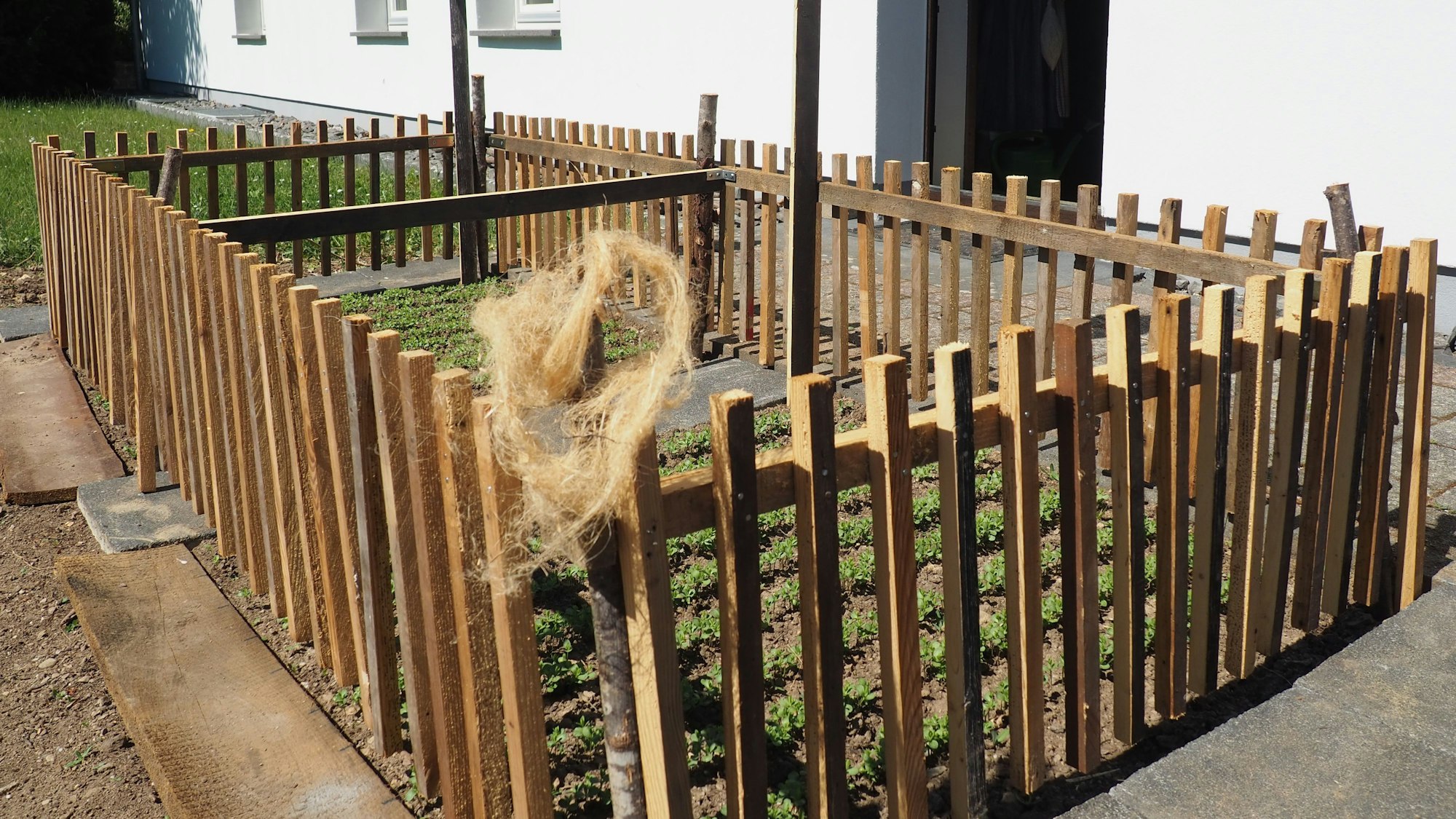 Ein Zaun steht um ein kleines Beet, in dem ein Schild steckt. An einem Zaunpfahl hängt ein Büschel Flachs-Fasern.