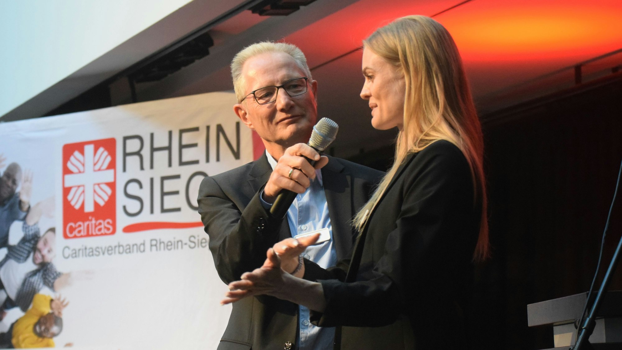 Ein Mann und eine Frau auf einer Veranstaltungsbühne. Im Hintergrund steht eine Werbetafel mit der Aufschrift Caritasverband Rhein-Sieg.