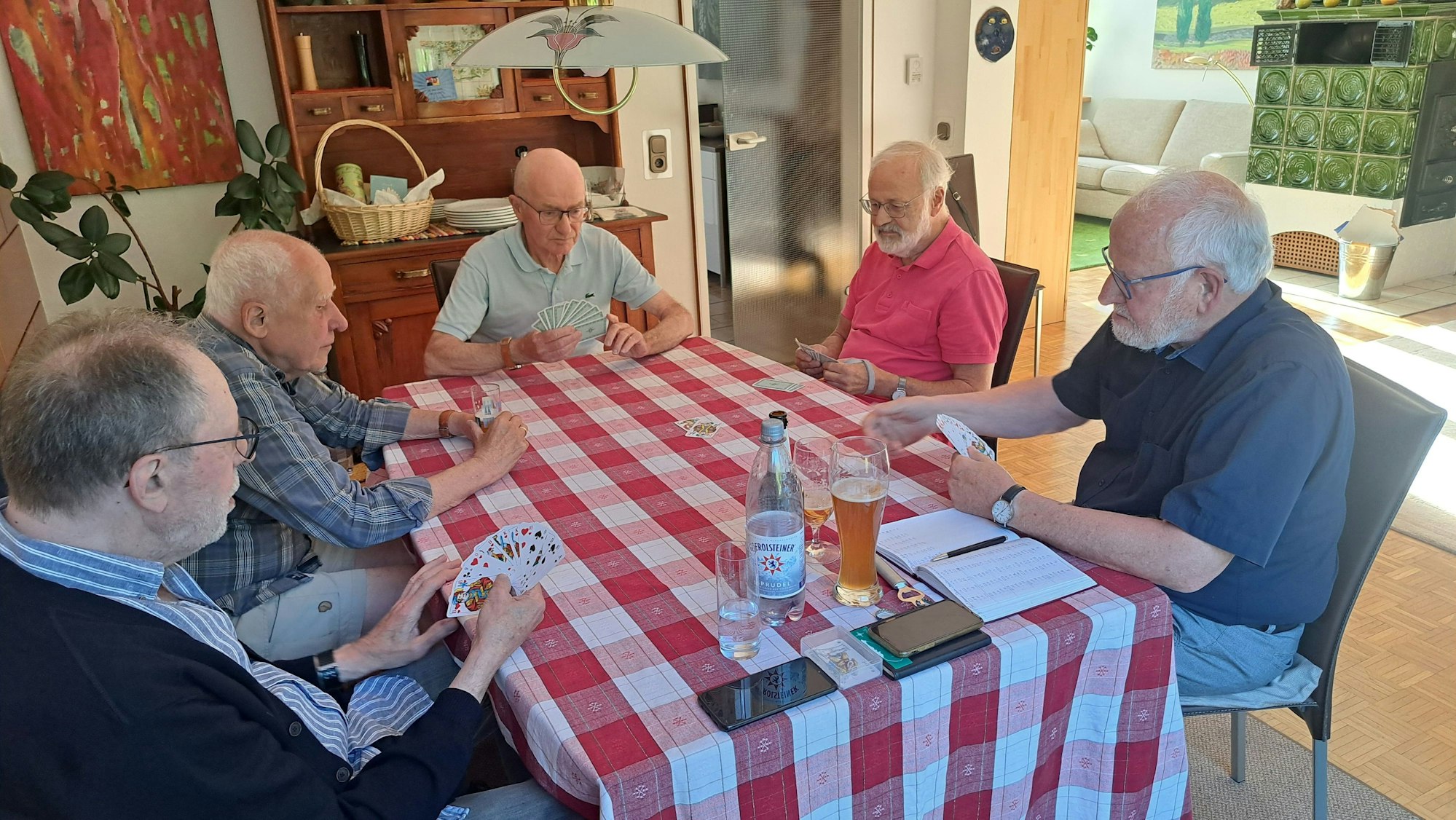 An einem Tisch mit rot-weißem Tischtuch sitzen fünf Männer, vier von ihnen schauen konzentriert auf die Karten in ihren Händen.