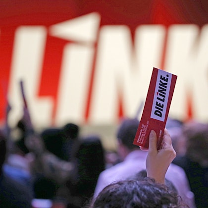 In einer Hand hält jemand, der nicht zu sehen ist, einen Zettel, auf dem „Die Linke“ steht.