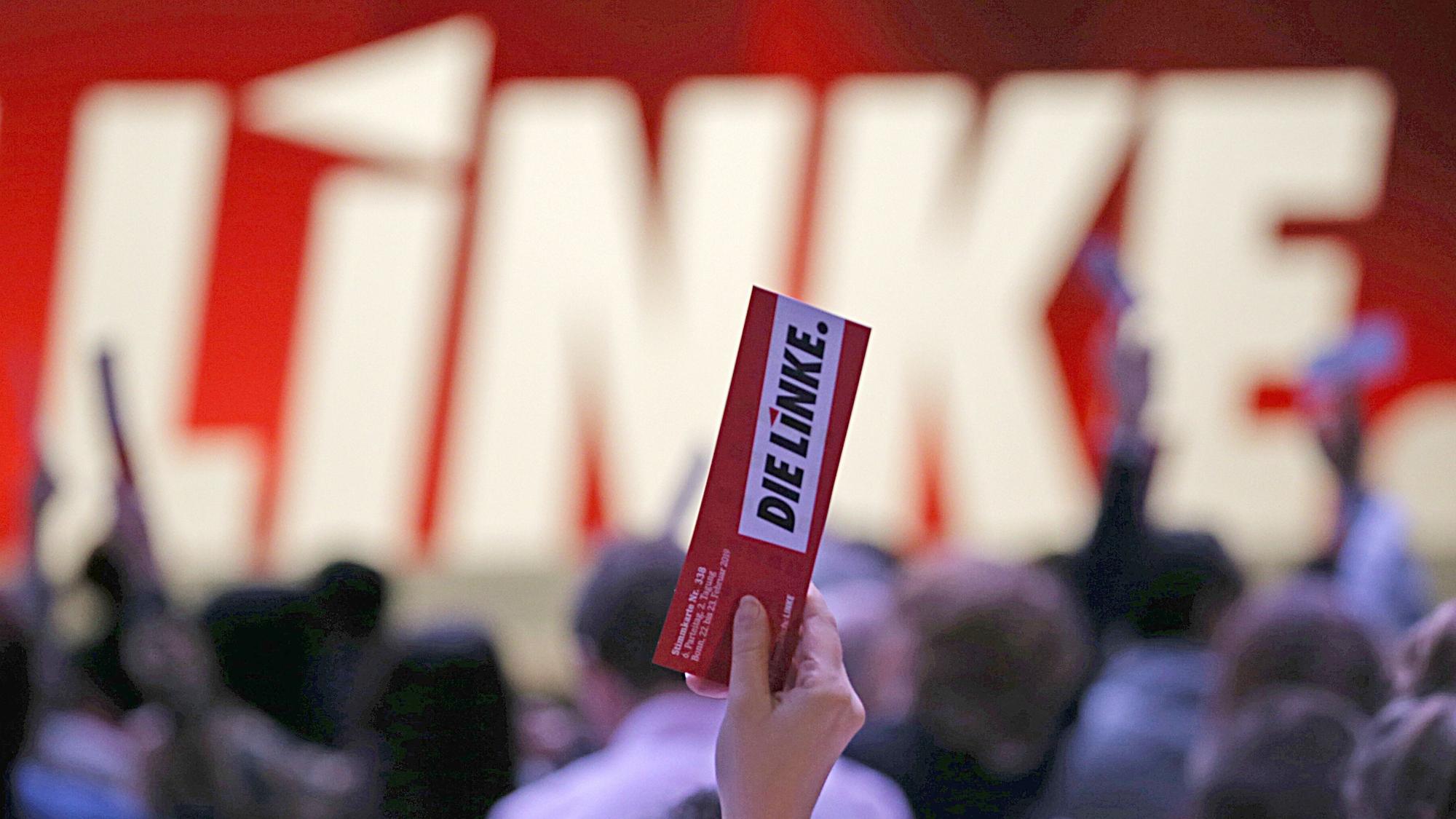 In einer Hand hält jemand, der nicht zu sehen ist, einen Zettel, auf dem „Die Linke“ steht.