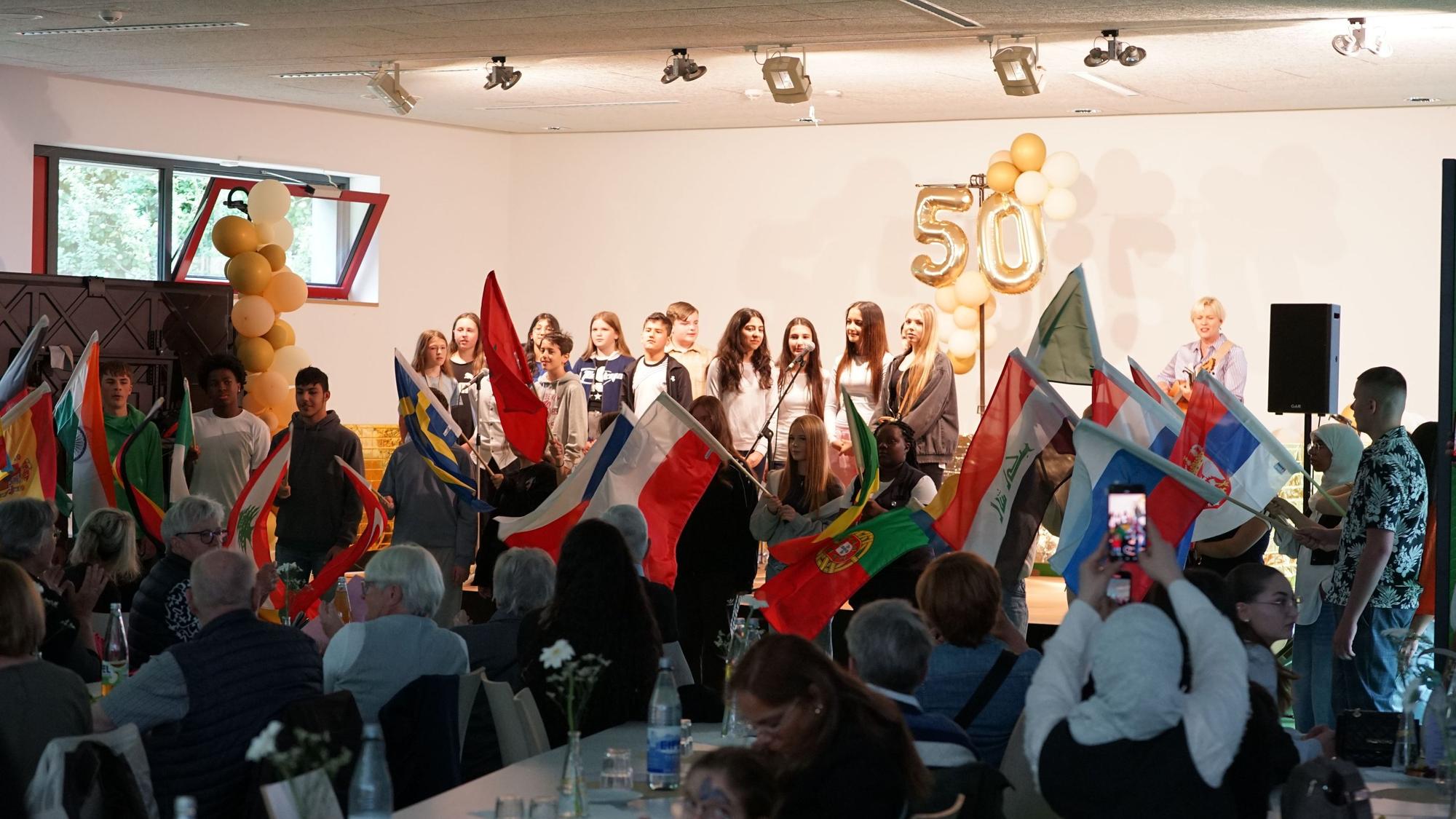 Auf dem Bild sind Kinder auf einer Bühne, internationale Fahnen und das Publikum zu sehen.