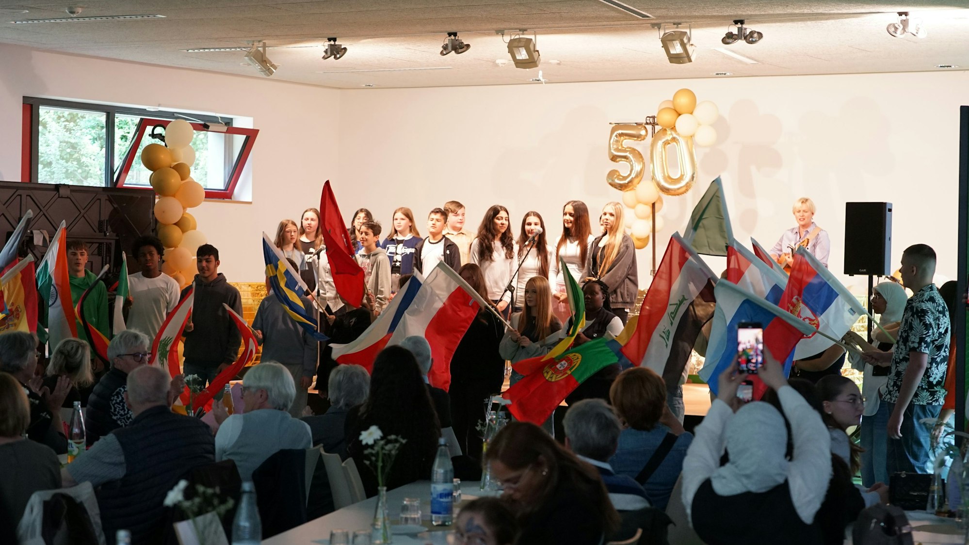 Auf dem Bild sind Kinder auf einer Bühne, internationale Fahnen und das Publikum zu sehen.