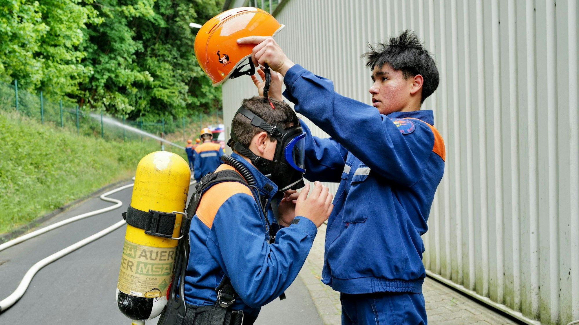 Ein junger Mann setzt einem anderen Feuerwehrkollegen in Vollmontur mit Sauerstoffflasche und Maske einen Schutzhelm auf.