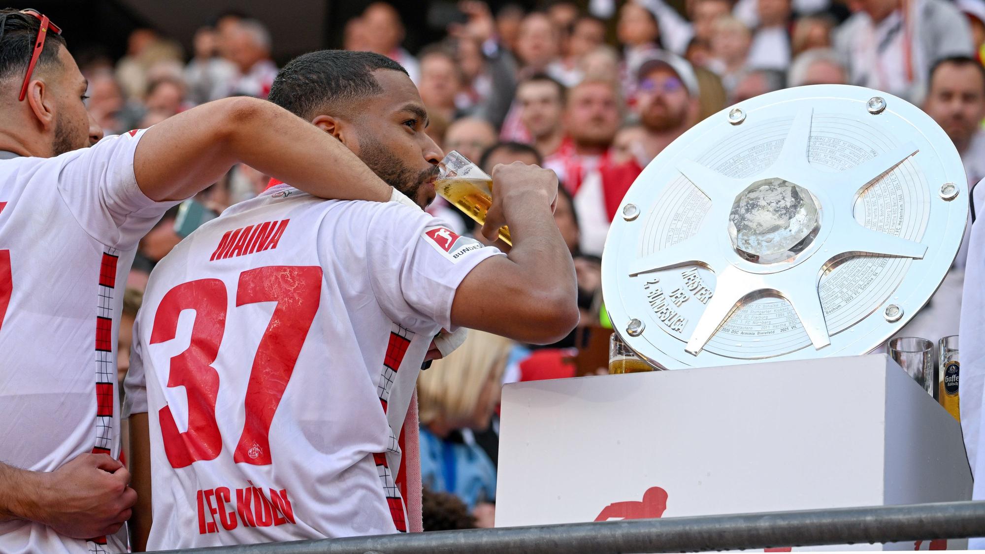 Kölns Linton Maina steht neben der Zweitliga-Meisterschale und feiert den Aufstieg mit einem Schluck Kölsch. Der Offensivspieler verkündete nach dem Schlusspfiff seine Vertragsverlängerung.