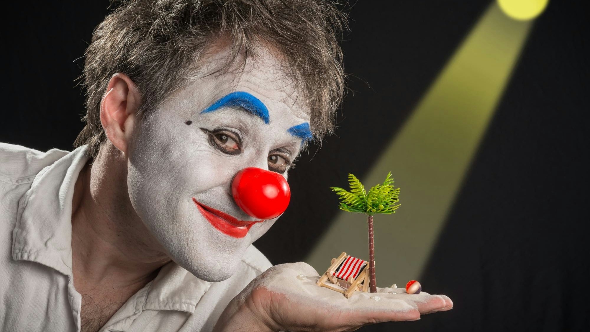 Clown Calvero ist geschminkt und hält eine Palme und einen Liegestuhl im Miniformat auf der Hand.